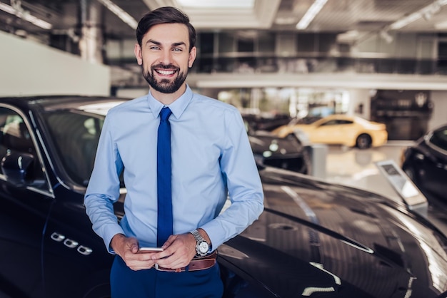 Retrato de vendedor sorridente parado no carro com smartphone no salão da concessionária