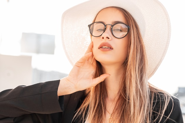 Retrato de verão ensolarado jovem bonito com belos lábios nos elegantes óculos com cabelo comprido, elegante jaqueta preta vintage chapéu de palha branco em dia ensolarado. Garota atraente e luz solar.