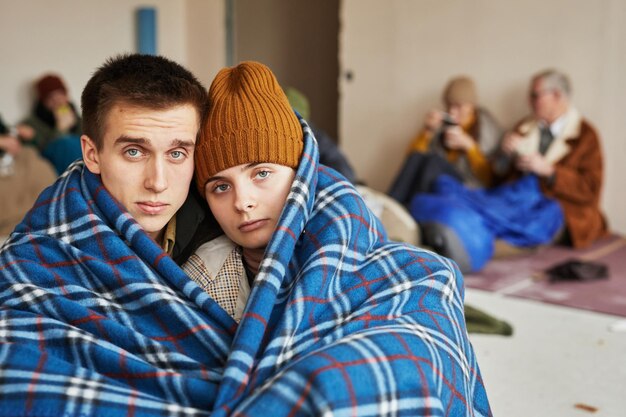 Retrato de vista frontal de um jovem casal caucasiano escondido em abrigo de refugiados coberto com cobertor no floo