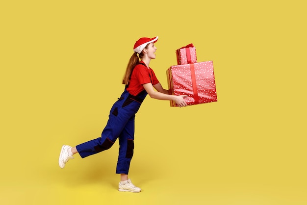 Retrato de vista lateral da mulher de correio sendo apressada para entregar caixas de presentes para clientes entrega porta a porta com vestindo macacão e boné vermelho Estúdio interno isolado em fundo amarelo