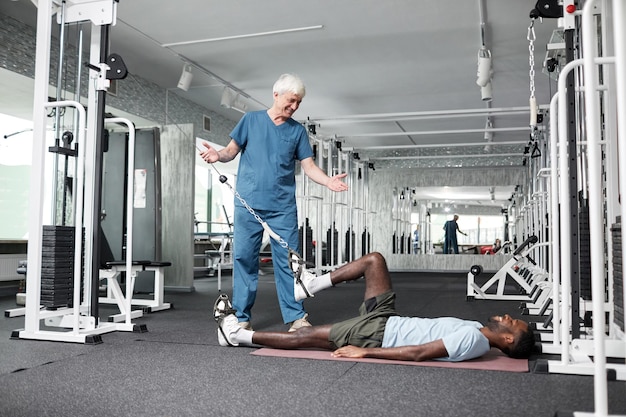 Retrato de vista lateral de especialista em fisioterapia ajudando jovem com exercícios de reabilitação em gy