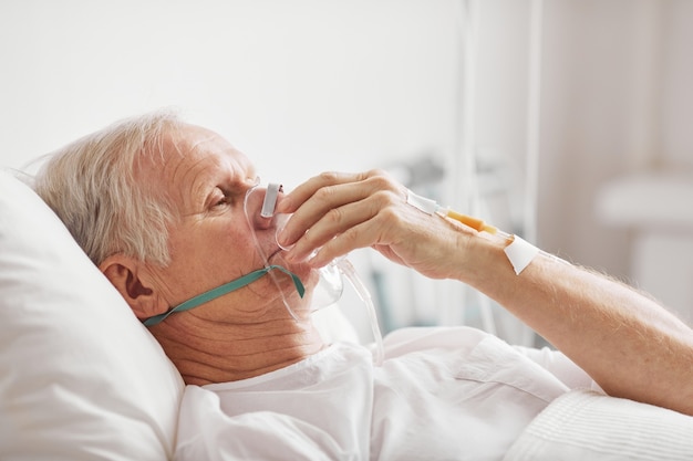 Retrato de vista lateral de homem idoso doente deitado em uma cama de hospital com máscara de suplementação de oxigênio e iv, copie o espaço