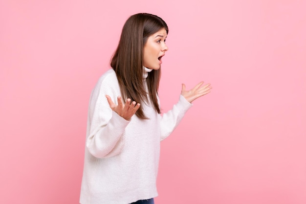 Retrato de vista lateral de jovem segurando as mãos em um gesto furioso furioso, gritando de raiva e raiva, vestindo suéter branco estilo casual. Tiro de estúdio interior isolado no fundo rosa.