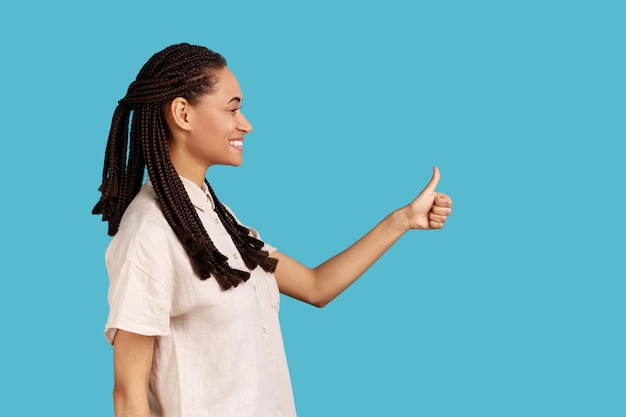 Retrato de vista lateral de mulher com dreadlocks pretos sorrindo alegremente, mantém os polegares para cima, aprova algo, tem expressão alegre, vestindo camisa branca. tiro de estúdio interior isolado sobre fundo azul.