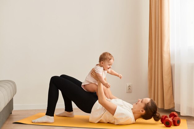 Retrato de vista lateral de mulher feliz vestindo camiseta branca e leggins pretas fazendo exercícios esportivos com bebê nas mãos deitada no tapete de ioga e fazendo exercícios para bunda