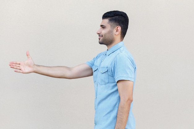 Retrato de vista lateral de perfil de homem barbudo jovem bonito feliz na camisa azul em pé e dando a mão ro saudação ou lidar com o sorriso. tiro de estúdio interior isolado no fundo da parede bege claro.