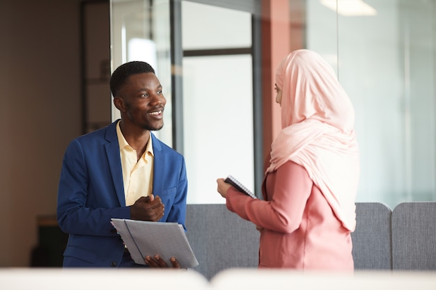 Retrato de vista lateral de um homem afro-americano sorridente, conversando com uma jovem empresária muçulmana e segurando documentos no interior de um escritório moderno, copie o espaço