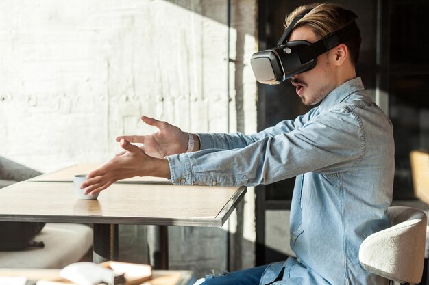 Retrato de vista lateral de um jovem adulto barbudo surpreso na camisa jeans azul sentado no café usando vr e assistindo vídeo no simulador com braços levantados e boca aberta Estilo de vida interior