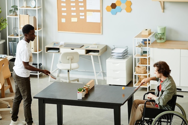 Foto retrato de vista lateral de um jovem com deficiência desfrutando de um jogo de tênis de mesa na sala de descanso do escritório a