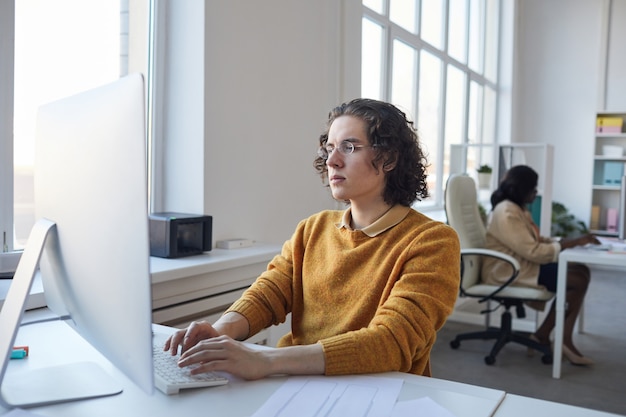 Retrato de vista lateral de um jovem desenvolvedor de software usando o computador enquanto trabalhava na mesa no interior branco do escritório, copie o espaço