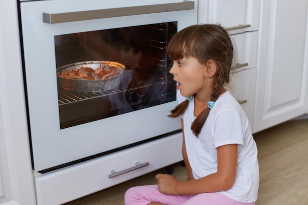 Retrato de vista lateral de uma garotinha atônita sentada perto do forno no chão na cozinha olhando para dentro com torta assada ou bolo esperando sobremesa deliciosa criança vestindo roupas casuais