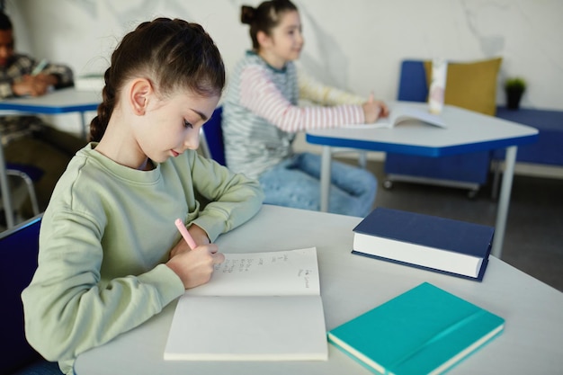 Retrato de vista lateral de uma jovem colegial escrevendo no caderno enquanto está sentado na mesa durante a cópia da aula