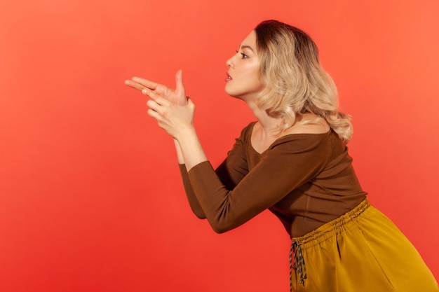 Retrato de vista lateral de uma mulher criminosa perigosa na blusa marrom apontando a arma do dedo ameaçando matar atirando com o gesto da arma bem no alvo Tiro de estúdio interno isolado em fundo vermelho