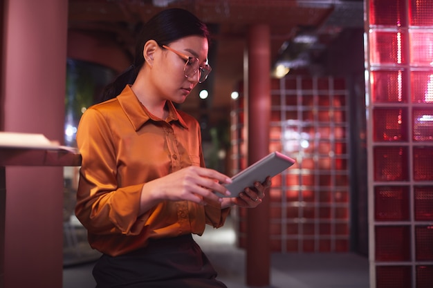 Retrato de vista lateral de uma mulher de negócios asiática contemporânea usando tablet digital enquanto trabalhava no interior de um escritório futurista