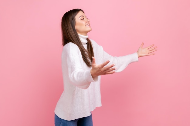 Retrato de vista lateral de uma mulher feliz, estendendo as mãos para abraçar, dando abraços grátis e dando boas-vindas, vestindo suéter branco estilo casual. Tiro de estúdio interior isolado no fundo rosa.