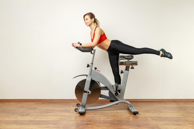 Foto retrato de vista lateral de uma mulher magra e atraente em pé na bicicleta ergométrica com treino de cardio de perna levantada vestindo meias esportivas e top vermelho estúdio interior tiro no fundo da parede cinza