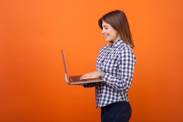 Retrato de vista lateral de uma mulher positiva ocupada com cabelo castanho em camisa casual quadriculada em pé digitando no laptop sorrindo satisfeito com o trabalho freelance estúdio interno tiro isolado em fundo laranja