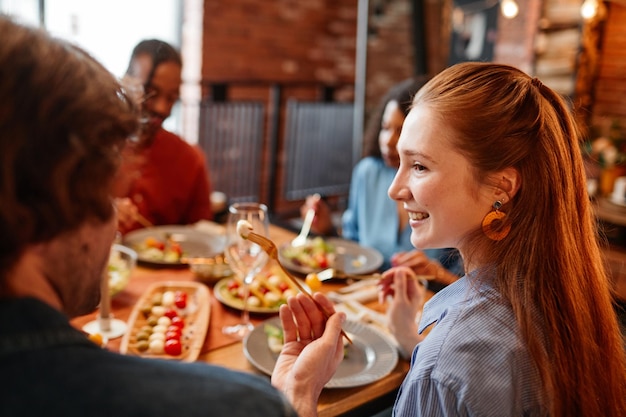 Retrato de vista lateral de uma mulher ruiva sorridente conversando com amigos enquanto desfruta de um jantar em coz