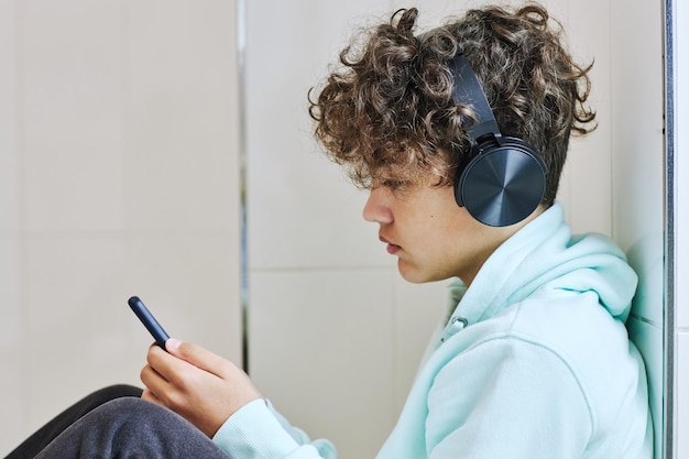 Retrato de vista lateral mínima de estudante adolescente sentado no chão e usando smartphone com fones de ouvido