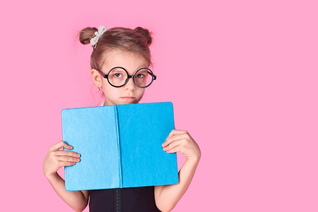 Retrato dela ela menina positiva alegre atraente atraente bonita sexy da escola que realiza nas mãos que escondem atrás da preparação aberta do exame do livro isolada sobre o fundo cor-de-rosa.