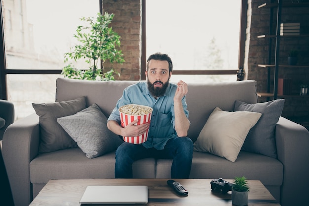 Retrato dele, ele é simpático, atraente, assustado, nervoso, moreno, sentado no divã, assistindo a uma série de TV, passando um tempo em um loft industrial, estilo moderno, interior, casa de quartos dentro de casa