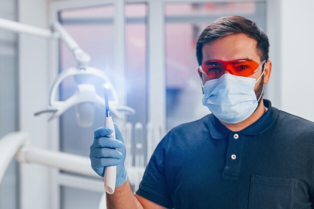 Retrato de dentista con gafas protectoras rojas y con herramienta en la mano que se encuentra en el interior de la clínica.