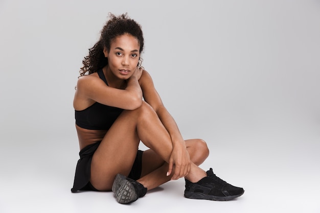 Retrato de una deportista africana bastante joven que se relaja después del entrenamiento aislado sobre la pared gris