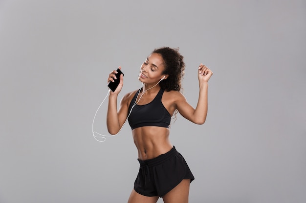 Retrato de una deportista alegre afroamericana aislada sobre fondo gris, escuchando música con auriculares, sosteniendo el teléfono móvil, bailando