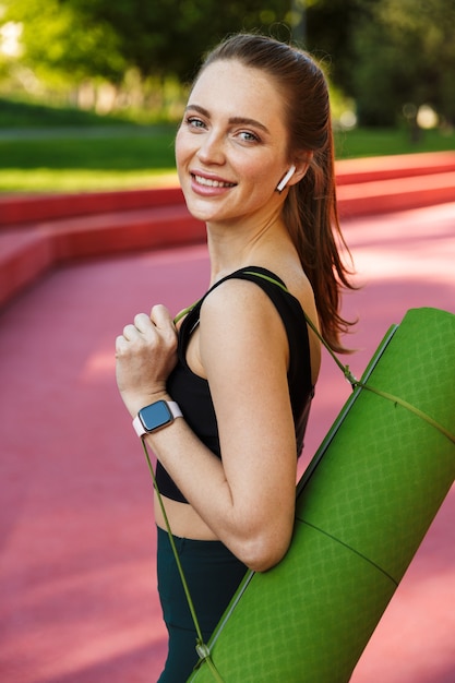 Retrato de deportista saludable vistiendo chándal y earpods llevando colchoneta de fitness durante el paseo por el parque de la ciudad
