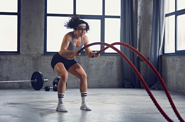 Retrato dinâmico de jovem mulher atlética em equipamento de ginástica fazendo exercícios de corda de batalha em movimento em