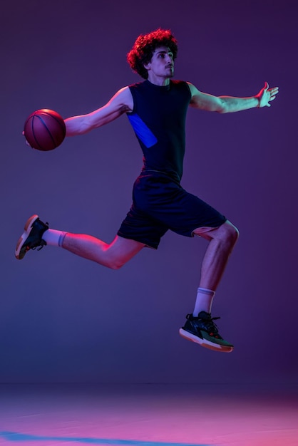 Retrato dinâmico do jovem jogador de basquete musculoso em treinamento de movimento isolado fundo roxo na luz neon
