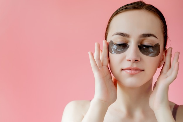 Retrato de disparo a la cabeza de cerca a mujer joven sonriente con máscara de parches hidratantes debajo de los ojos mirando a cámara disfrutando del procedimiento de cuidado de la piel.