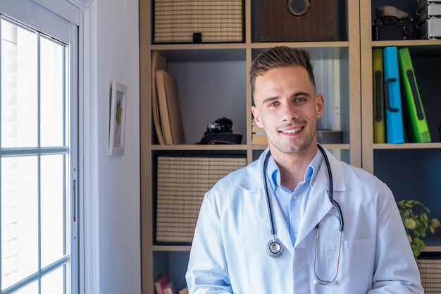 Retrato de disparo en la cabeza de sonriente joven médico de cabecera o médico con uniforme médico blanco y gafas mirando a la cámara posando en el hospital, feliz médico caucásico muestra habilidades de confianza en el lugar de trabajo de la clínica