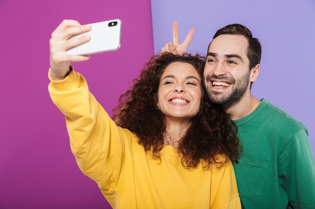 Foto retrato de divertida pareja caucásica en ropa colorida divirtiéndose mientras toma selfie foto en teléfono celular aislado