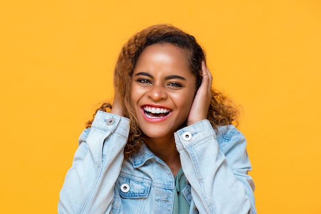 Retrato divertido de una joven afroamericana feliz riéndose mientras se cubre los oídos con ambas manos en un estudio aislado de fondo amarillo