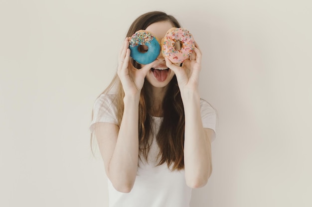 Retrato divertido de una mujer sosteniendo donas azules y rosas cerca de sus ojos y mostrando una lengua