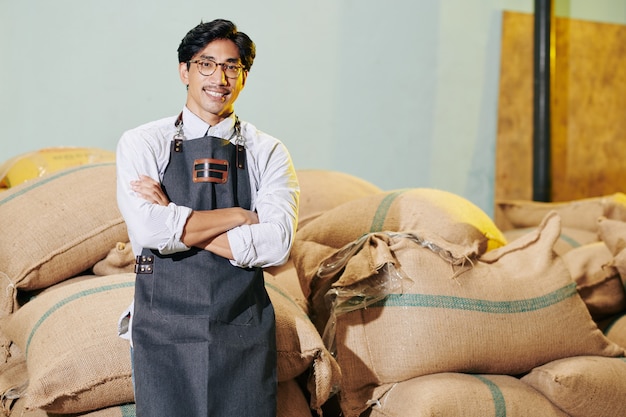 Retrato do alegre proprietário de um torreão vietnamita no avental, cruzando os braços e olhando para a câmera diante de sacos de grãos de café