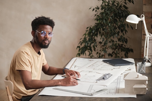 Foto retrato do arquiteto afro-americano criativo de conteúdo em óculos redondos, sentado à mesa com plantas no escritório
