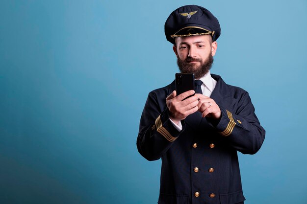 Retrato do capitão do avião segurando mensagens de texto para smartphone com tripulantes remotos em estúdio com fundo azul. Piloto com uniforme de aviação, assistindo vídeo online no celular, plano médio