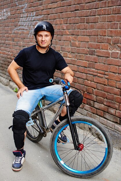 Retrato do cara de capacete em uma bicicleta sentado contra a parede de tijolo vermelho