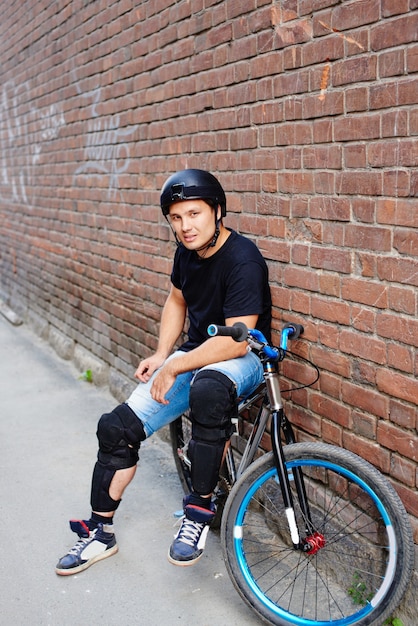 Retrato do cara de capacete em uma bicicleta sentado contra a parede de tijolo vermelho