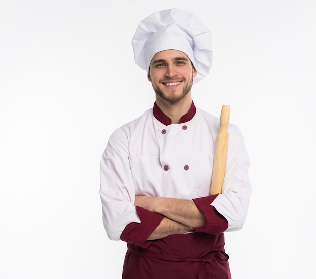 Retrato do chef dentuço positivo cozinheiro com roupa boina, branca, tendo ferramentas nos braços cruzados, olhando para a câmera isolada no fundo branco.