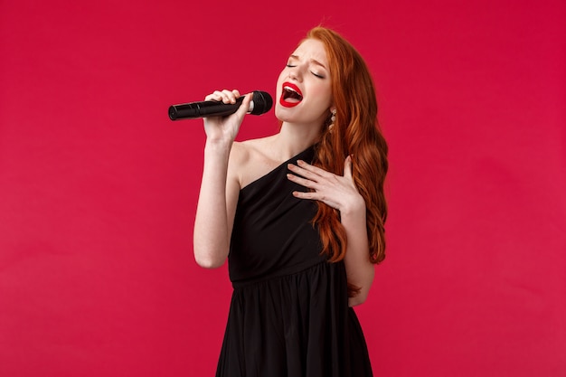 Retrato do close-up da cantora linda apaixonada executar músicas, vestido preto, fechar os olhos e mostrando seus sentimentos através da música, segure o microfone, assistir karaokê na noite das meninas