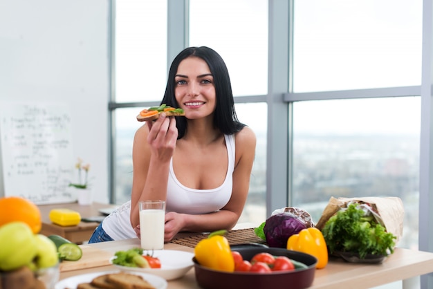 Retrato do close-up da mulher em pé na cozinha, apoiando-se na mesa de madeira, lanche. Menina sorridente mantém estilo de vida saudável.