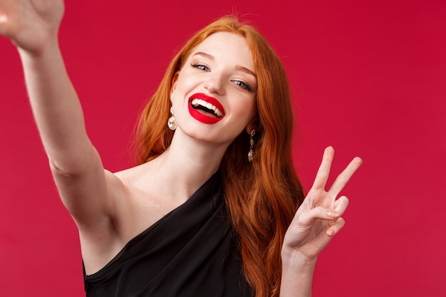 Retrato do close-up da mulher ruiva encantadora feliz curtindo a festa, tomando selfie com sinal de paz e um sorriso radiante, use vestido de noite preto e batom vermelho, fique em uma parede vermelha