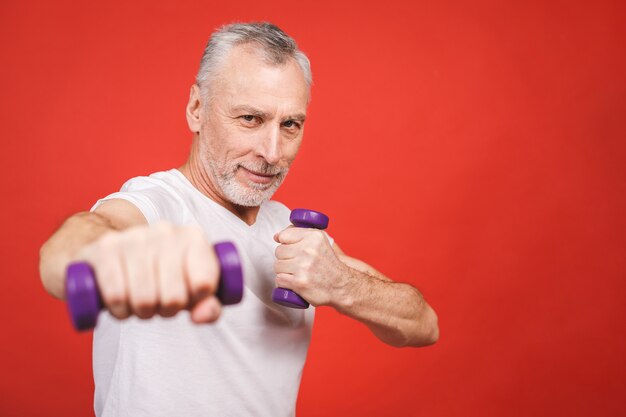 Retrato do close-up de um homem sênior exercitar com halteres contra fundo vermelho