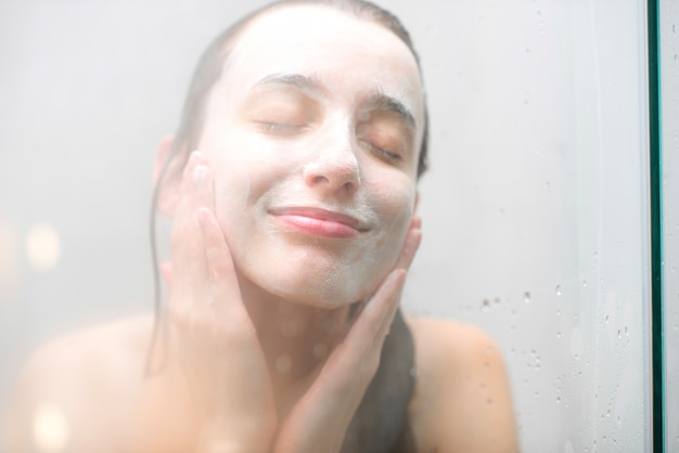 Retrato do close-up de uma mulher com sabonete no rosto molhado, em pé atrás do vidro do chuveiro. Imagem com foco suave
