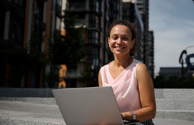 Retrato do close-up de uma mulher de negócios fofo, de etnia mista, trabalhando no laptop sentado na escadaria da cidade e sorrindo, olhando para a câmera. Negócios, trabalho remoto distante e conceito de start-up