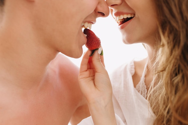 Retrato do close-up do lindo casal alegre apaixonado, comendo saboroso morango juntos em fundo branco. Menina sorridente com longos cabelos castanhos claros alimentando seu namorado sorridente com frutas frescas