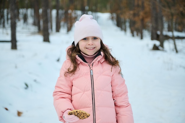 Retrato do comprimento da cintura de criança feliz encantadora menina caucasiana de 5 anos de idade agradável lanches ao ar livre enquanto caminhava em uma floresta coberta de neve desfrutando de lazer de inverno em um lindo dia ensolarado de neve